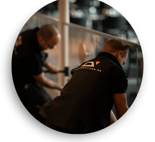 Kaksi asentajaa mustissa työvaatteissa asentaa lasikaidetta julkisessa tilassa. He työskentelevät huolellisesti lasin säätämisen ja kiinnityksen parissa. Lasi heijastaa tilaa ja tuo avoimuutta ympäristöön.