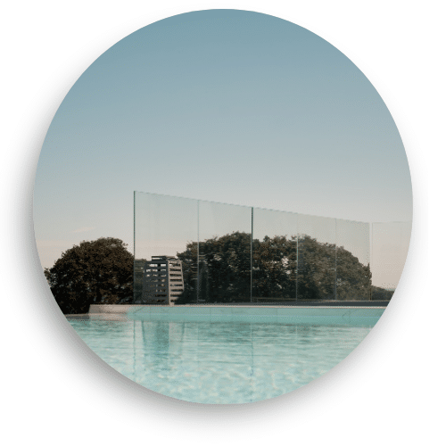 Tolpaton kirkas lasikaide asennettuna uima-altaan reunalle. Suuret lasipaneelit kiinnitettynä huomaamattomaan alaprofiliin, tarjoten esteettömän näkymän maisemaan. Vesi heijastaa taivasta rauhallisessa ympäristössä.