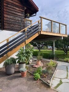 Korkealle sijoitettu terassi, jossa puinen kaide ja kirkas lasi talon hirsiseinää vasten. Näkymä avautuu puutarhaan ja vehreyteen.