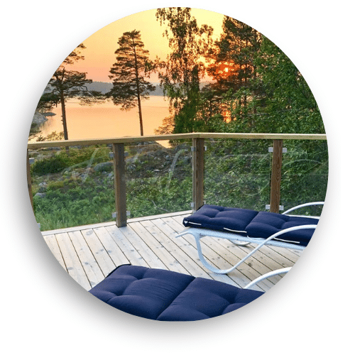 Puuterassille asennettu lasikaide, jossa puutolpat ja klämfästit kehystävät selkeitä lasipaneeleja. Näköala järvelle ilta-auringossa. Kaksi tummasävyistä aurinkotuolia terassilla.