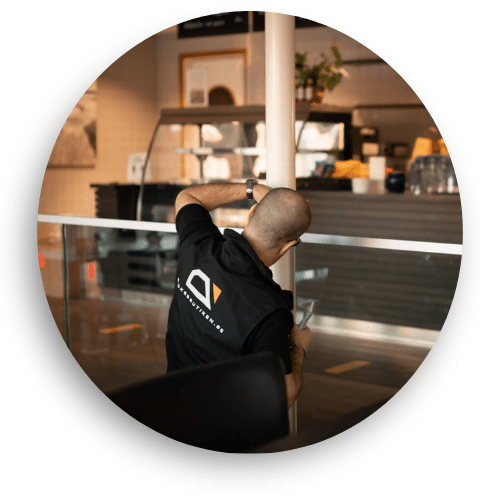 Asentaja säätämässä lasikaidetta julkisessa kahvilassa. Kaide koostuu kirkkaasta lasista ja ruostumattomista yksityiskohdista, tyylikäs ja toimiva ratkaisu turvallisuusluokituksella