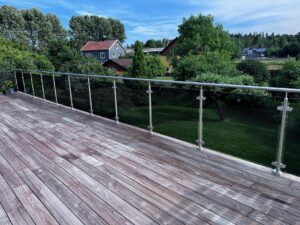 Parvekekaide savulasi- ja ruostumattomilla tolpilla puuterassilla, näkymä puutarhaan ja taloon.