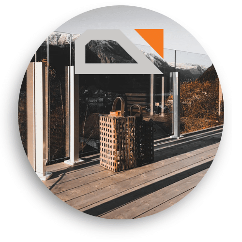 Lasikaide valkoisilla tolpilla ja klämfästeillä asennettuna puuterassille. Läpinäkyvät lasipaneelit kehystävät näkymän vuoristoon ja laaksoon. Kaksi koristeellista lyhtyä seisoo terassilla.