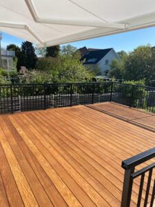 Musta taottu kaide puuterassin ympärillä, markiisi, aurinkoinen laudoitus ja rehevä vihreys taustalla.