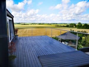 Ruostumattomasta teräksestä valmistettu vaijerikaide tummanharmaalla puulattialla terassilla. Esteetön näkymä pelloille ja vihreille niityille.