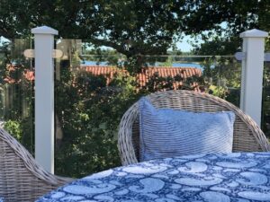 Lähikuva terassista valkoisella alumiinikaiteella ja kirkkaalla lasilla, rottinkikalusteet, siniset tekstiilit ja järvimaisema taustalla.