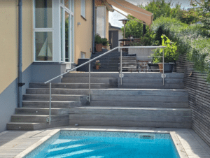 Ruostumattomasta teräksestä valmistettu kaide leveän puuportaan vieressä uima-altaalla – täydentää lasiaidan ja tarjoaa yhtenäisen suojan.