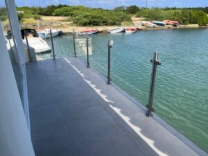 Lasikaide ruostumattomilla tolpilla laiturin varrella, näkymä venesatamaan ja vehreyteen taustalla