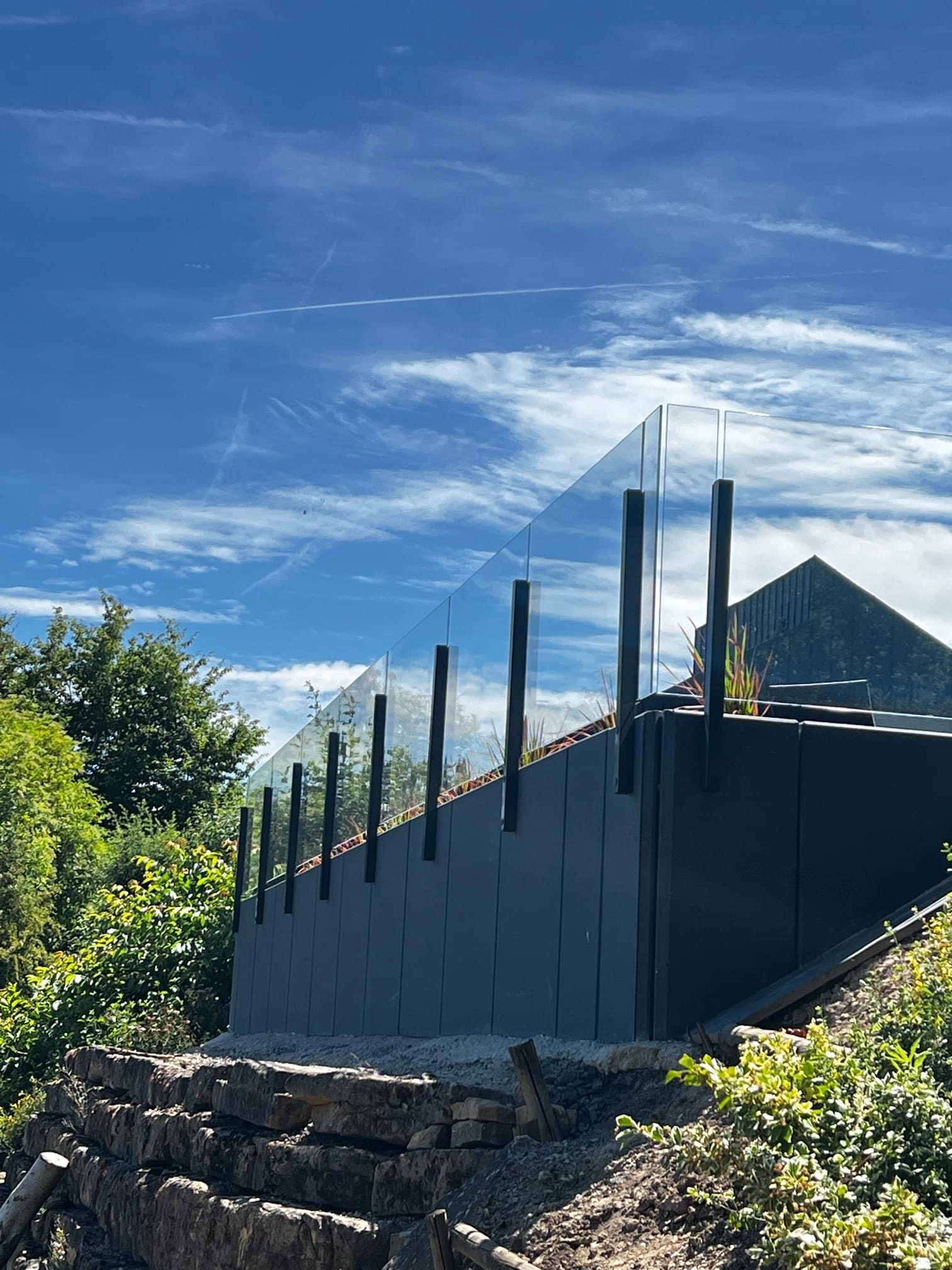 Tyylikäs lasikaide korkeilla mustilla alumiinitolpilla, korotettu terassi tummalla verhouksella ja vehreällä ympäristöllä.
