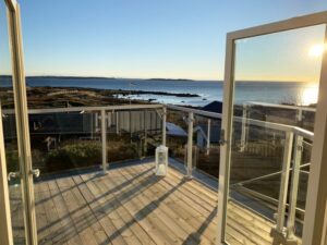 Parveke lasikaiteella ja valkoisella alumiinirungolla. Avoimet pariovet ja taustalla auringonlasku meren ja saariston yllä.