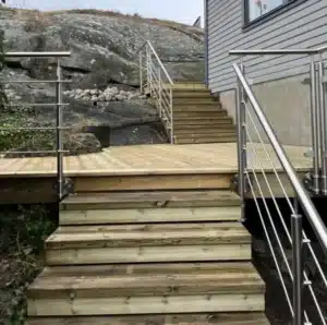 Puuverhoiltu terassi ja portaat, joissa on ruostumaton kaide vaakasuuntaisilla säleillä. Kaide seuraa sekä portaita että tasanteita. Talon julkisivu on harmaata paneelia, ja taustalla näkyy kallioinen maisema.